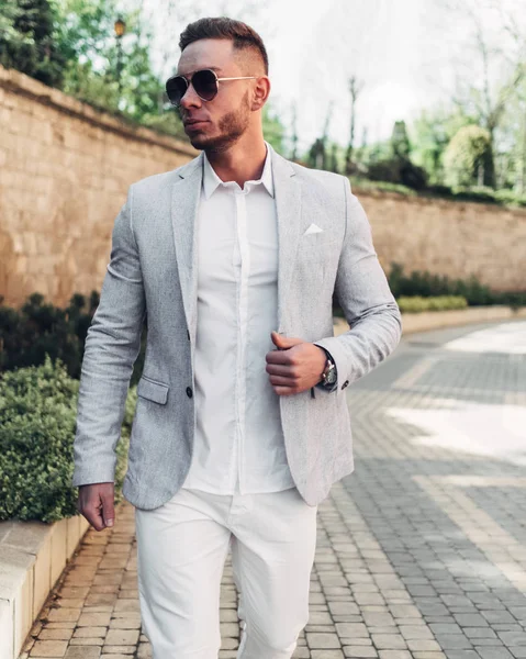 Sakallı ve güneş ışığı altında sokakta şık takım elbise poz içinde gözlüklü genç adam