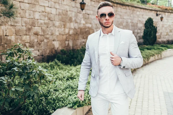 Sakallı ve güneş ışığı altında sokakta şık takım elbise poz içinde gözlüklü genç adam