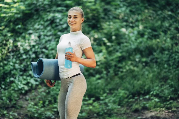 Fitness Woman Preparing for Yoga in the Early Morning Park. Wearing Stylish Sport Outfit. Blue Bottle of Water and Yoga Mat. Healthy Lifestyle.