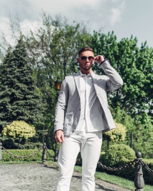 Sakallı ve güneş ışığı altında sokakta şık takım elbise poz içinde gözlüklü genç adam