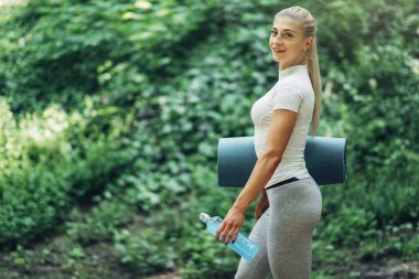 Fitness kadın erken sabah parkta Yoga için hazırlanıyor. Şık spor kıyafeti giymiş. Mavi şişe su ve Yoga Mat. Sağlıklı yaşam tarzı.