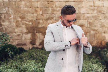 Sakallı ve güneş ışığı altında sokakta şık takım elbise poz içinde gözlüklü genç adam