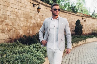 Sakallı ve güneş ışığı altında sokakta şık takım elbise poz içinde gözlüklü genç adam
