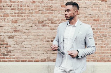 Sakallı ve güneş ışığı altında sokakta şık takım elbise poz içinde gözlüklü genç adam