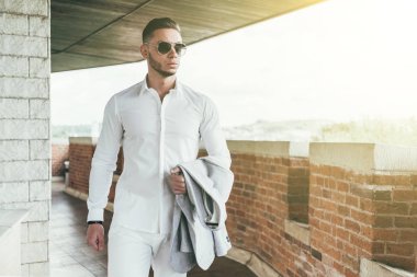 Sakallı ve güneş ışığı altında sokakta şık takım elbise poz içinde gözlüklü genç adam