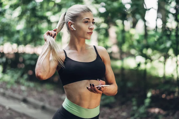 Woman Runner in the Summer Morning Park Listening to Music on Smartphone Using Bluetooth Earphones. Female Fitness Girl Jogging on Path Outside.