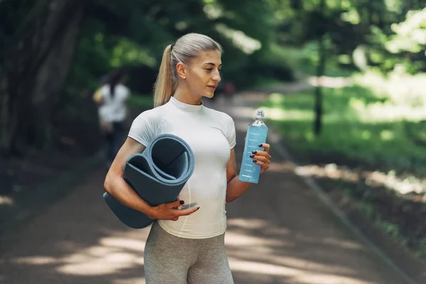 Fitness kadın erken sabah parkta Yoga için hazırlanıyor. Şık spor kıyafeti giymiş. Mavi şişe su ve Yoga Mat. Sağlıklı yaşam tarzı.