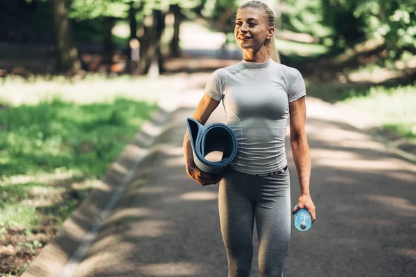 Fitness Woman Preparing for Yoga in the Early Morning Park. Wearing Stylish Sport Outfit. Blue Bottle of Water and Yoga Mat. Healthy Lifestyle.