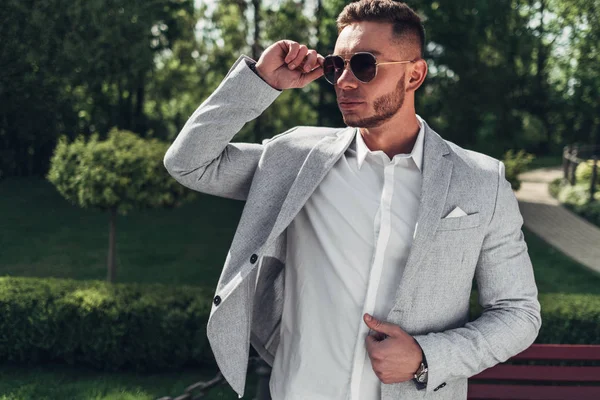 Sakallı ve güneş ışığı altında sokakta şık takım elbise poz içinde gözlüklü genç adam