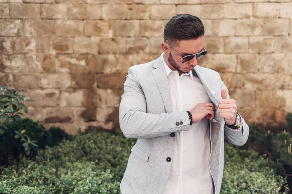 Sakallı ve güneş ışığı altında sokakta şık takım elbise poz içinde gözlüklü genç adam