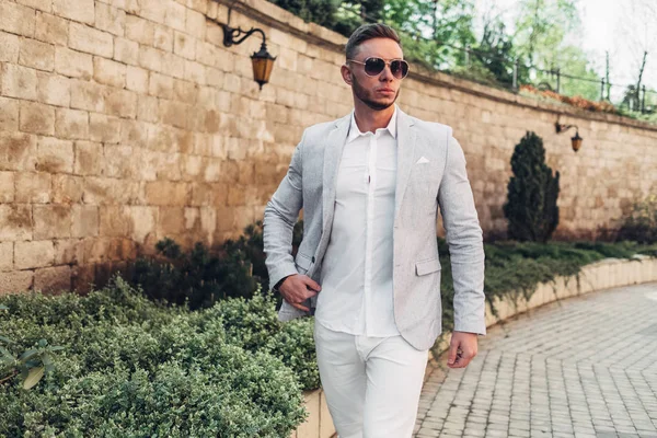 Sakallı ve güneş ışığı altında sokakta şık takım elbise poz içinde gözlüklü genç adam