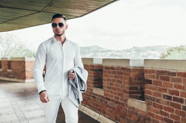 Sakallı ve güneş ışığı altında sokakta şık takım elbise poz içinde gözlüklü genç adam