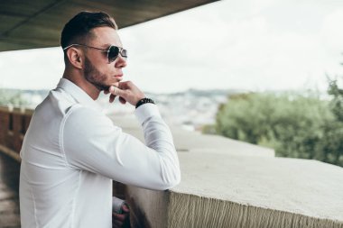 Sakallı ve güneş ışığı altında sokakta şık takım elbise poz içinde gözlüklü genç adam