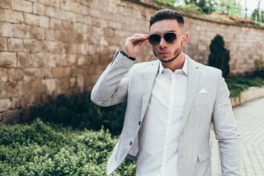 Sakallı ve güneş ışığı altında sokakta şık takım elbise poz içinde gözlüklü genç adam
