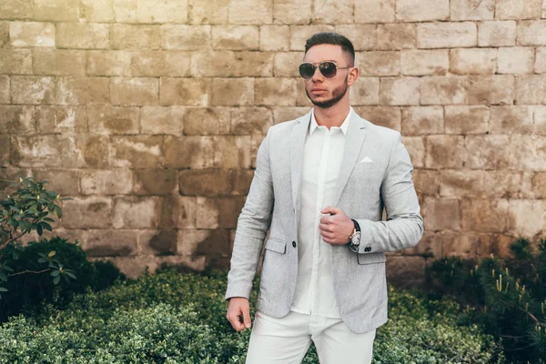 Sakallı ve güneş ışığı altında sokakta şık takım elbise poz içinde gözlüklü genç adam