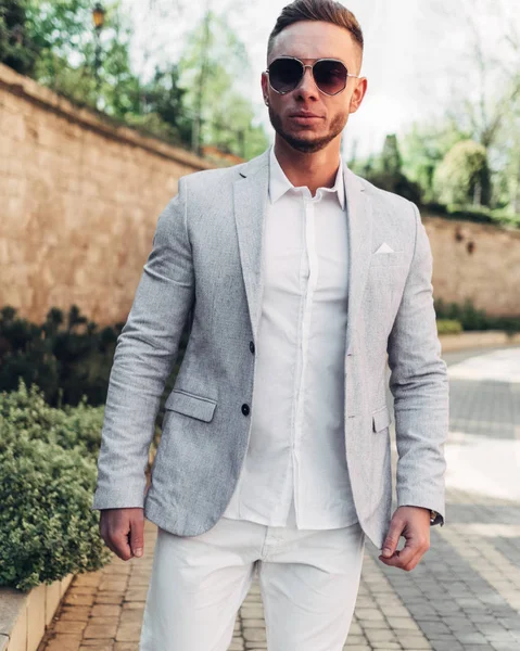Sakallı ve güneş ışığı altında sokakta şık takım elbise poz içinde gözlüklü genç adam