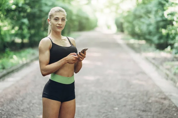 Woman Runner in the Summer Morning Park Listening to Music on Smartphone Using Bluetooth Earphones. Female Fitness Girl Jogging on Path Outside.