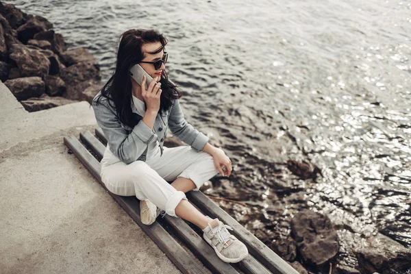 Όμορφη μελαχρινή κορίτσι στα γυαλιά ηλίου λευκό παντελόνι και τζιν μπουφάν κοντά στη λίμνη — Φωτογραφία Αρχείου