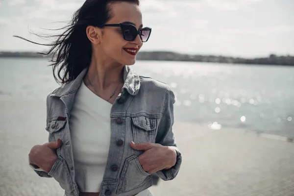 Vacker brunett flicka i solglasögon vita byxor och jeans jacka nära sjön — Stockfoto