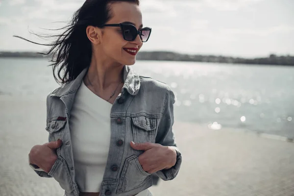 Hermosa chica morena en gafas de sol pantalones blancos y Jeans chaqueta cerca del lago —  Fotos de Stock