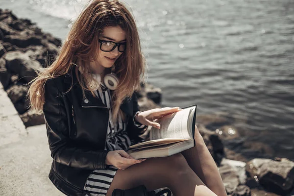 Retrato de verão ao ar livre da menina bonita nova em jaqueta de couro preto e óculos lê o livro perto do lago — Fotografia de Stock