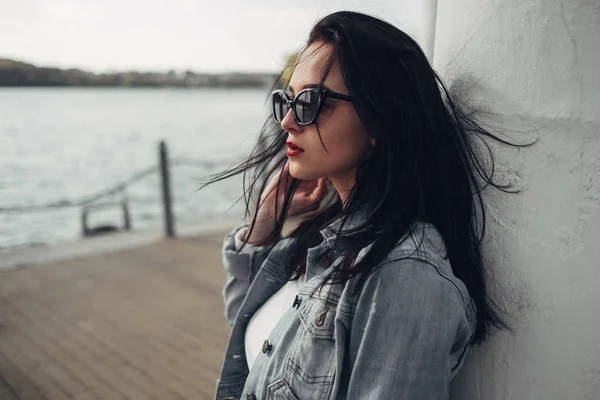 Vacker brunett flicka i solglasögon vita byxor och jeans jacka nära sjön — Stockfoto