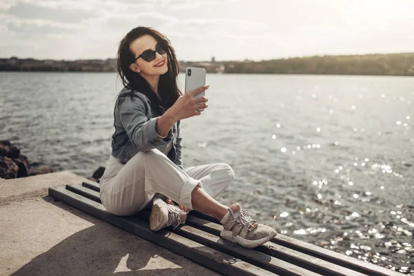 Красива брюнетка дівчина в сонцезахисних окулярах Білі штани і джинси куртка біля озера — стокове фото