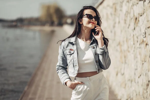 Piękna brunetka dziewczyna w okulary przeciwsłoneczne białe spodnie i dżinsy kurtka w pobliżu jeziora — Zdjęcie stockowe