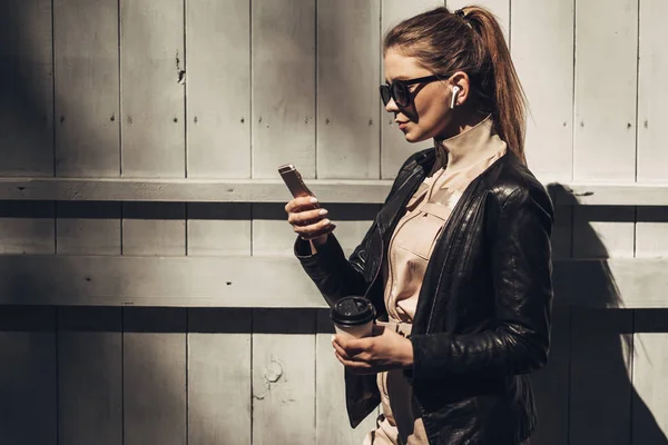 Portret stylowej dziewczyny nosi kobiet kombinezon i czarna skórzana kurtka pić kawę, korzystanie Smartphone — Zdjęcie stockowe