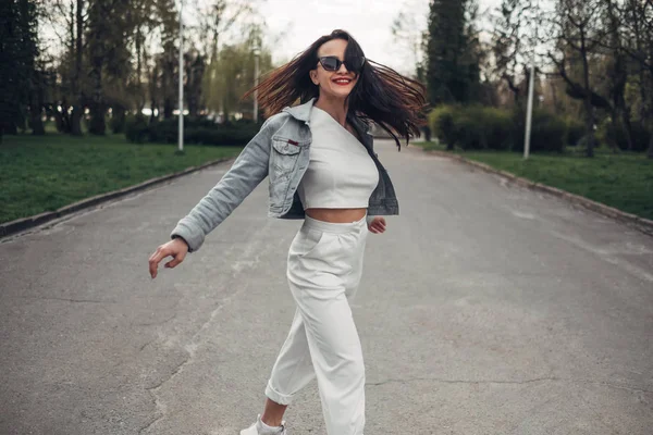 Schöne brünette Mädchen mit Sonnenbrille, weißer Hose und Jeansjacke, rotem Lippenstift — Stockfoto