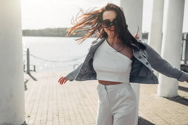 Belle fille brune en lunettes de soleil pantalon blanc et veste de jeans près du lac — Photo