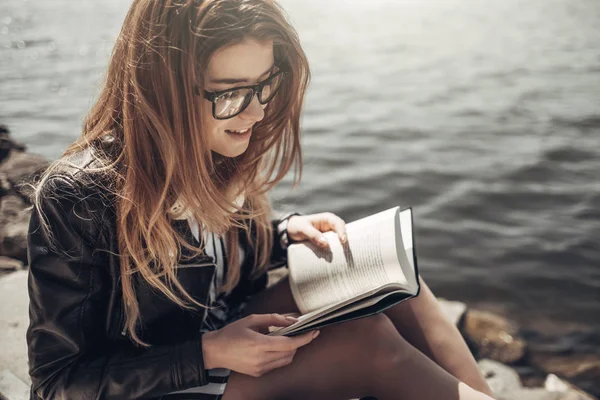 Retrato de verão ao ar livre da menina bonita nova em jaqueta de couro preto e óculos lê o livro perto do lago — Fotografia de Stock