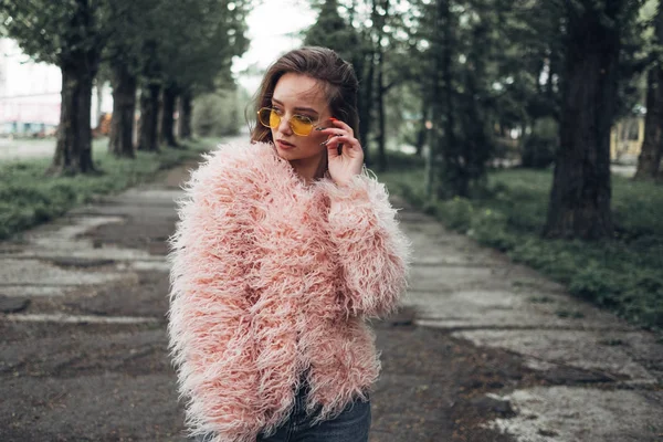 Porträtt av en snygg flicka i rosa päls och moderna gula solglasögon poserar på gatan — Stockfoto