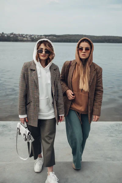 Retrato de duas meninas da forma, melhores amigos ao ar livre, vestindo jaqueta elegante, andando perto do lago — Fotografia de Stock