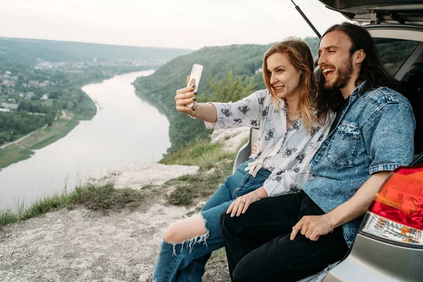 Young Stylish Traveling Couple Having Fun Sitting in Modern Car and Making Selfie, Travel and Road Trip Concept