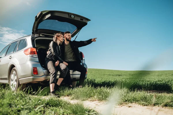 緑のフィールドでロードトリップで楽しい時間を過ごしている黒い革とジーンズのジャケットのように服を着た幸せな若い旅行者のカップル — ストック写真