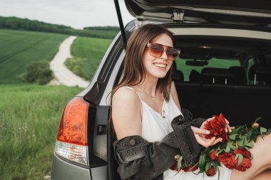 Beyaz Elbiseli Genç Tarz Kadını ve Kırmızı Şakayık Buketli Denim Ceketi Yeşil Tarladaki SUV Arabasının Bagajında Oturuyor