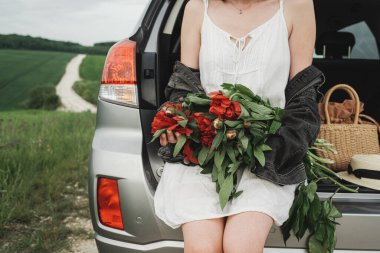 Beyaz Elbiseli Genç Tarz Kadını ve Kırmızı Şakayık Buketli Denim Ceketi Yeşil Tarladaki SUV Arabasının Bagajında Oturuyor