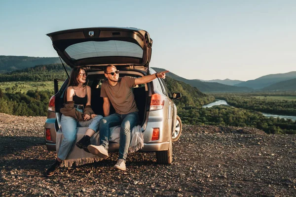 Young Traveler Couple on Road Trip, Man and Woman Sitting on the Opened Trunk of Their Car Over Sunset