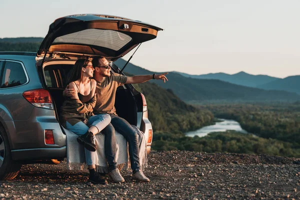 Young Traveler Couple on Road Trip, Man and Woman Sitting on the Opened Trunk of Their Car Over Sunset