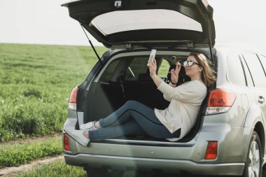 Açık Araba Bagajında Oturan ve Video Çağrı için Akıllı Telefon kullanan Genç Kadın