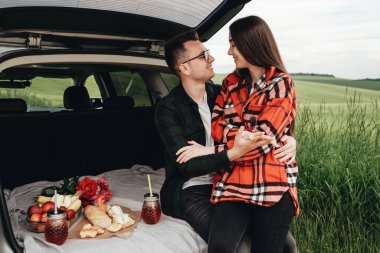 Araba bagajında oturan ve yolculuğun tadını çıkaran genç ve güzel bir çift, piknik konsepti.
