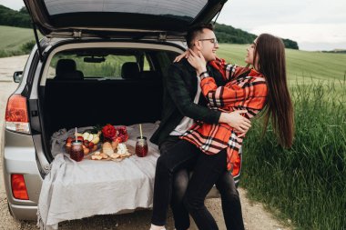 Araba bagajında oturan ve yolculuğun tadını çıkaran genç ve güzel bir çift, piknik konsepti.