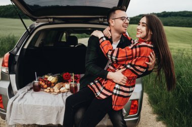 Araba bagajında oturan ve yolculuğun tadını çıkaran genç ve güzel bir çift, piknik konsepti.