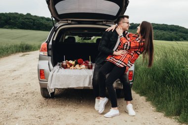Araba bagajında oturan ve yolculuğun tadını çıkaran genç ve güzel bir çift, piknik konsepti.