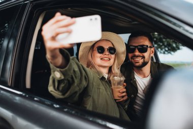 Kadın ve erkek arabada oturuyor, soğuk kahve içiyor ve akıllı telefonda selfie çekiyor, macera yolculuğunun tadını çıkarıyorlar.