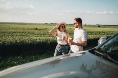 Erkek ve kadın arabanın yanında eğleniyor, soğuk kahve içiyor ve macera yolculuğunun tadını çıkarıyor.