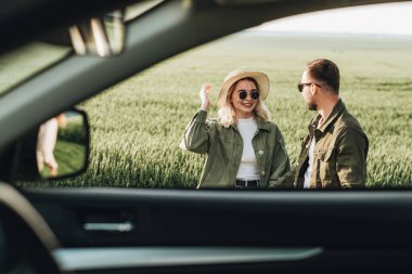 Erkek ve kadın zeytin ceketli giyinmiş, arabanın yanında eğleniyor, macera yolculuğunun tadını çıkarıyorlar.