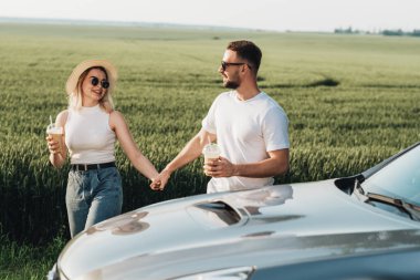 Erkek ve kadın arabanın yanında eğleniyor, soğuk kahve içiyor ve macera yolculuğunun tadını çıkarıyor.