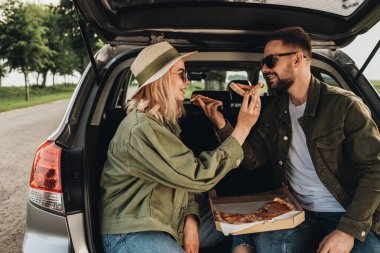 Kadın ve erkek, bir araba bagajında oturup pizza yiyor.